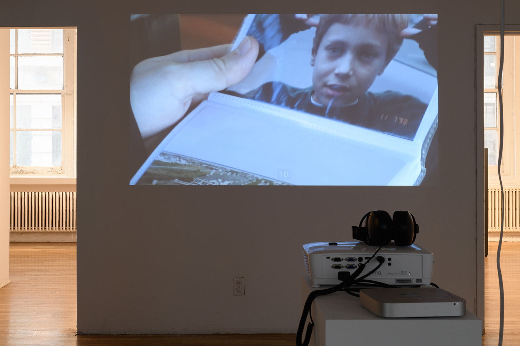 [A view of a color video projected at large-scale. The projected image, which shows a white hand holding a photo album open to a photo of a young person, who also appears to be white, wearing a blue sweater. On either side of the projected image are doorways leading to additional galleries.]